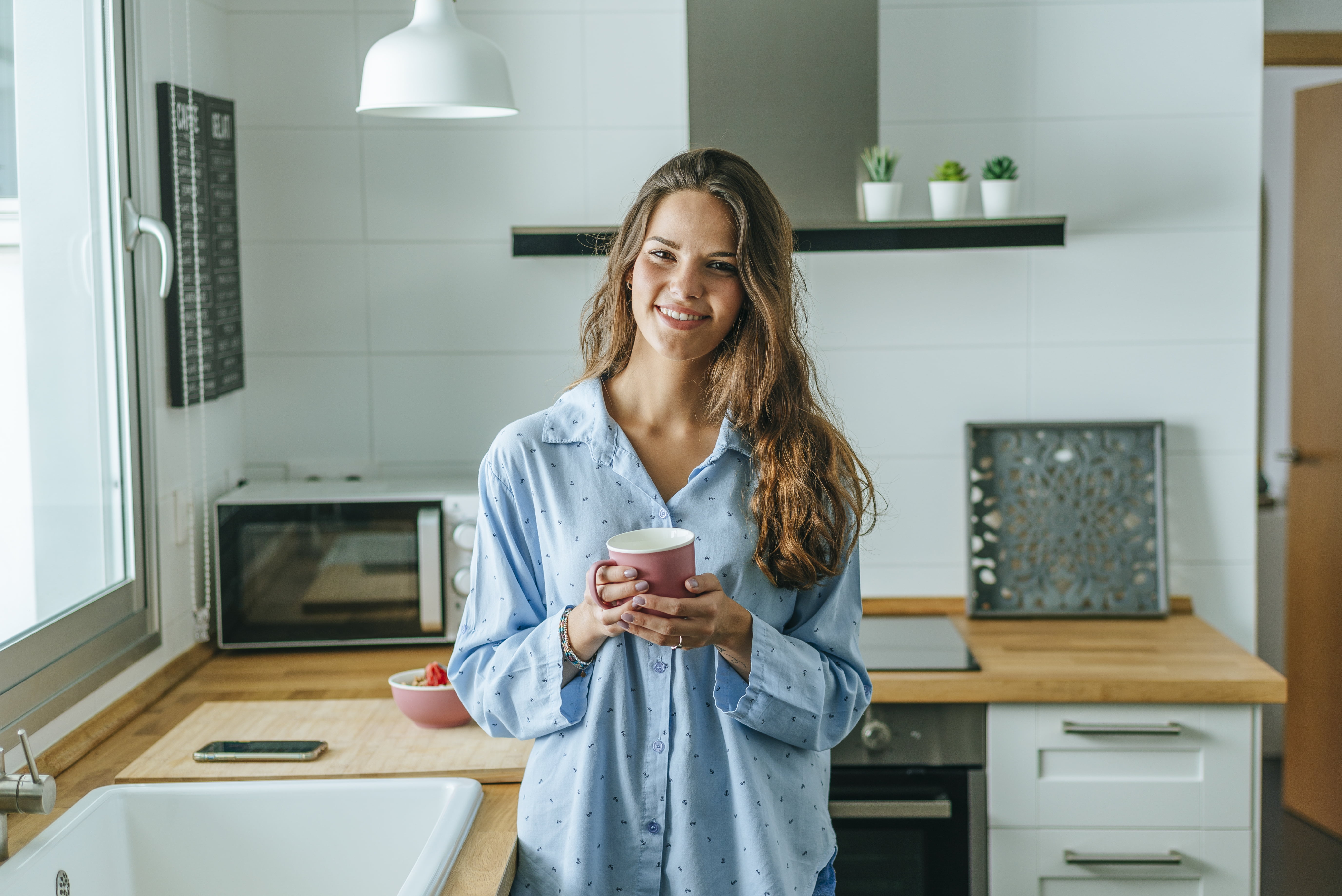 En kvinne står smilende på kjøkkenet med en kaffekopp i hånden